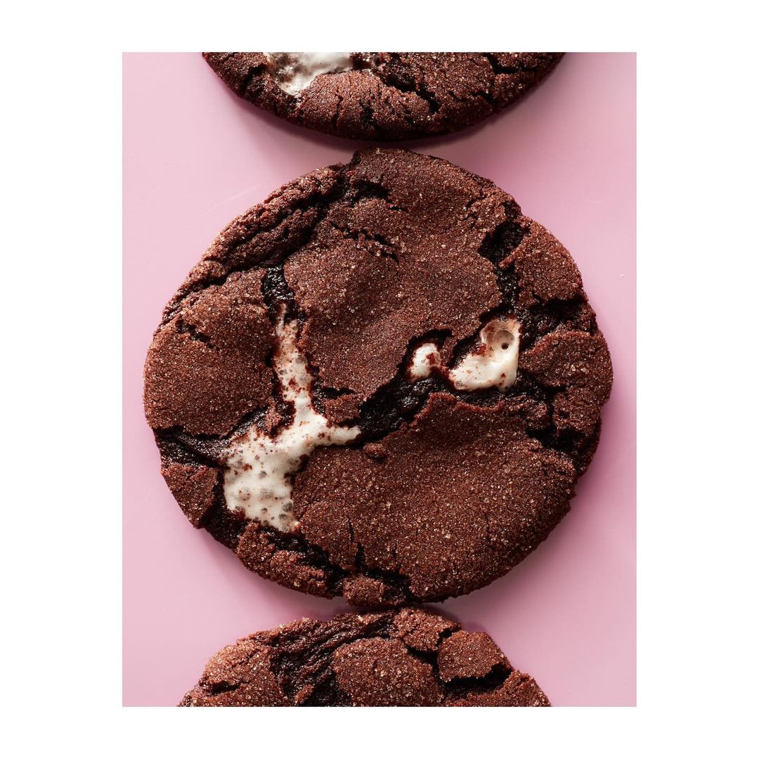 Overhead shot of three chocolate cookies with melted marshmallows on a light pink surface, highlighting texture and delicious details.