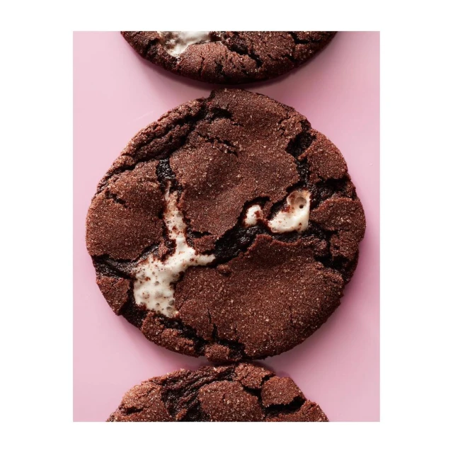 Overhead shot of three chocolate cookies with melted marshmallows on a light pink surface, highlighting texture and delicious details.