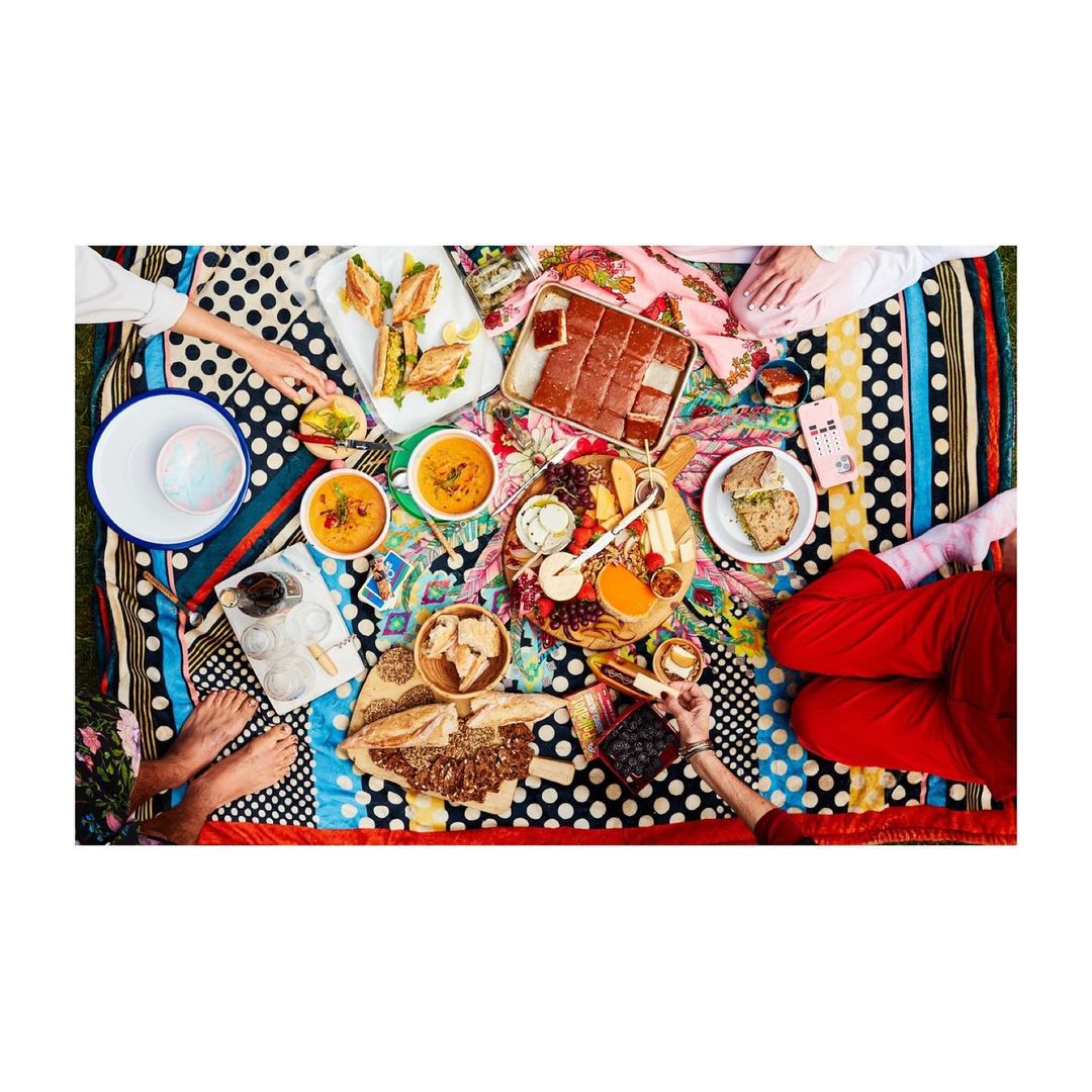 Overhead shot of a picnic spread with assorted food platters, bowls, and drinks on a colorful patterned blanket with hands visible.