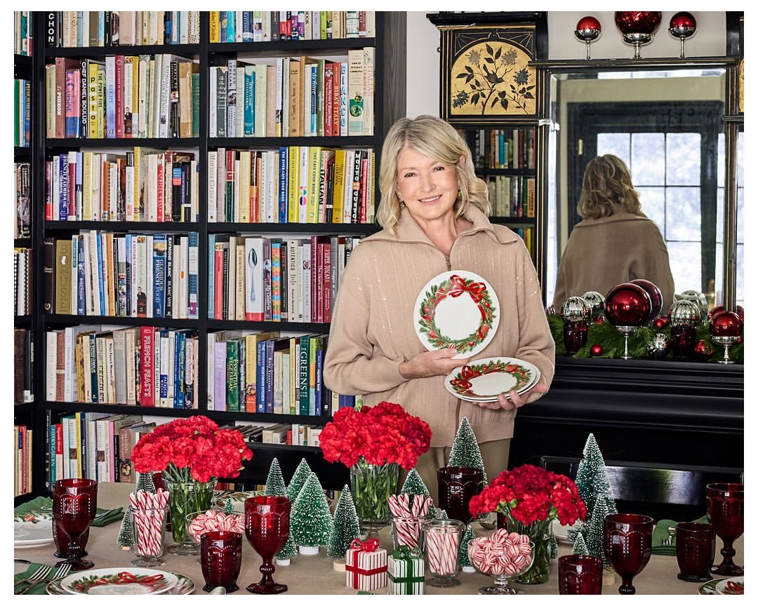 Mature woman holding holiday plates at a dining table, surrounded by flower arrangements, holiday decorations and book shelves in background.