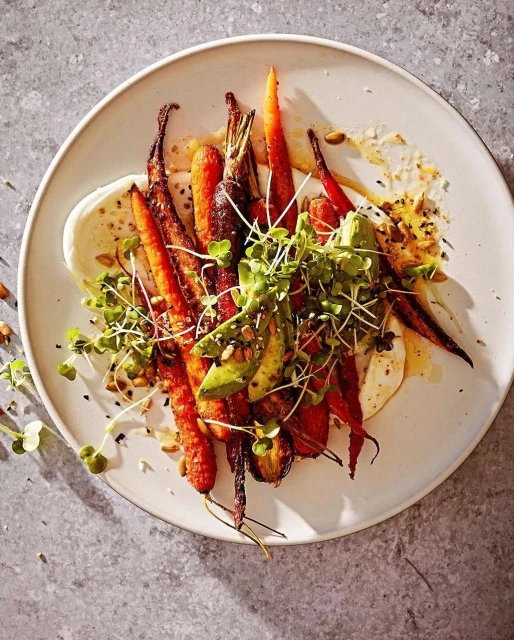 Overhead shot of a white plate featuring roasted carrots with toppings such as sliced avocado and sprouts on a gray background.