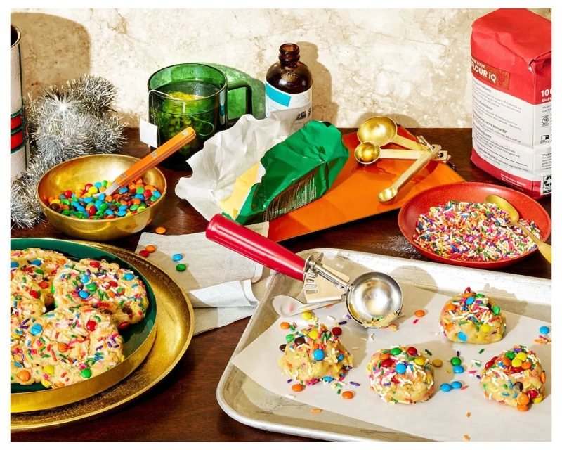 Festive holiday baking scene with cookies on a tray, sprinkles, M&Ms, flour, butter, and various baking tools on a wooden table.