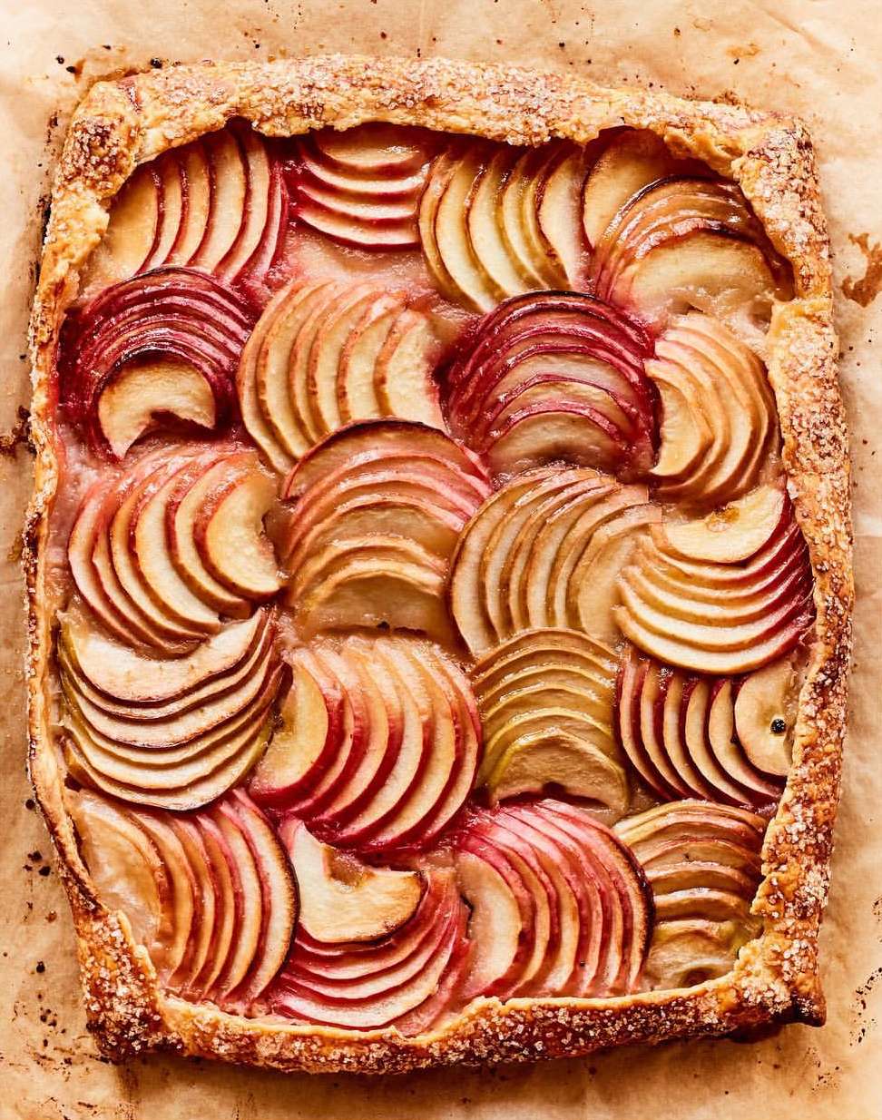 Overhead shot of a rectangular apple tart with sliced apples arranged in concentric curves on flaky crust, sitting on parchment paper.