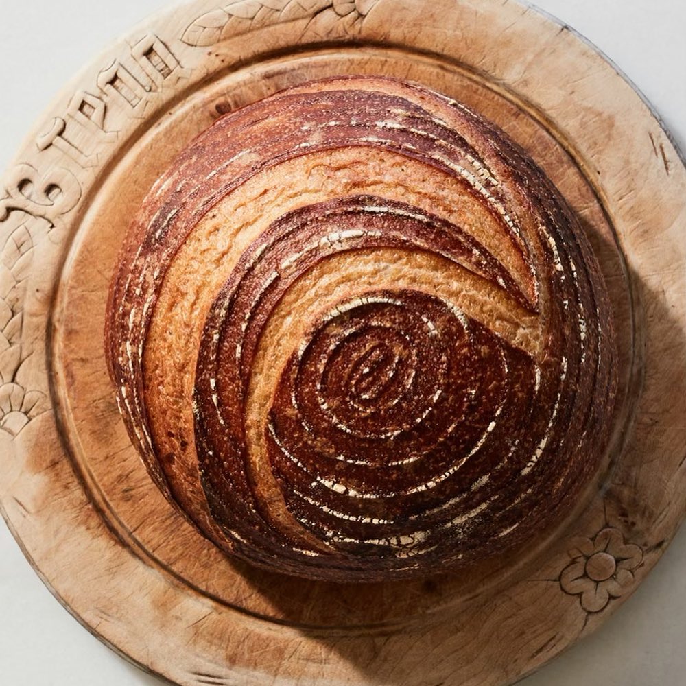 Overhead shot of a round loaf of sourdough bread on a carved wooden board, showcasing its crust and spiral scoring pattern.