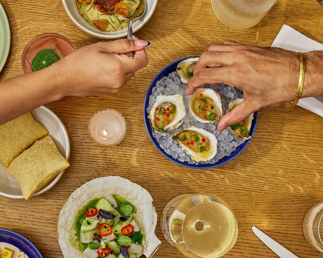 Overhead shot of table with oysters, finger foods, drinks, and two people preparing to eat at a restaurant or home dining table.