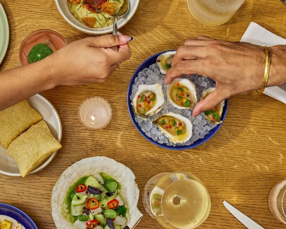 Overhead shot of table with oysters, finger foods, drinks, and two people preparing to eat at a restaurant or home dining table.