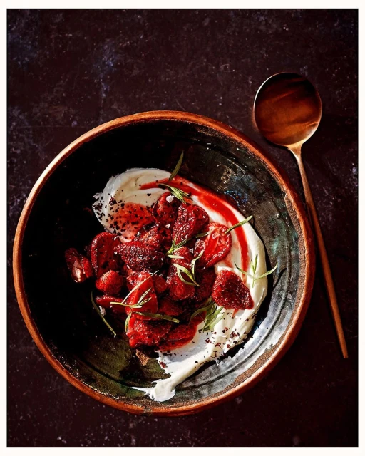 Overhead shot of a rustic bowl containing strawberries, cream, herbs, and red sauce, with a spoon on a dark surface.
