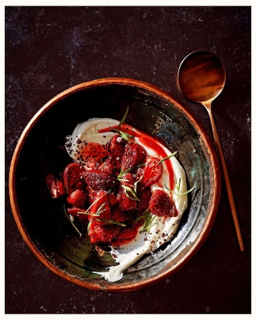 Overhead shot of a rustic bowl containing strawberries, cream, herbs, and red sauce, with a spoon on a dark surface.