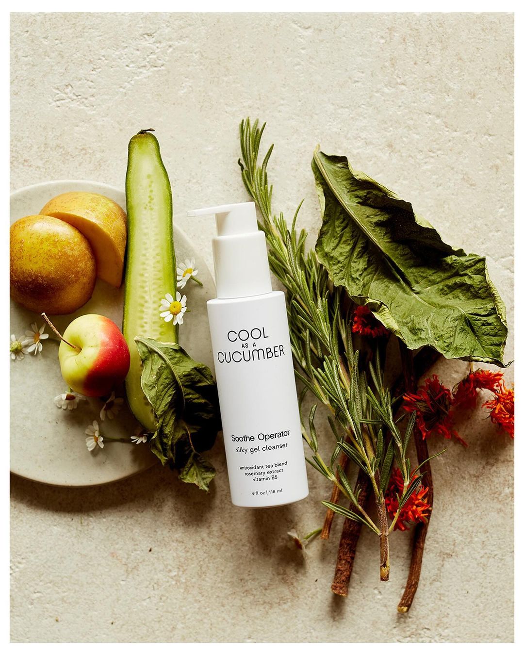 Still life of skincare product bottle with cucumber, rosemary sprigs, apple, and sliced pears on an off-white surface.