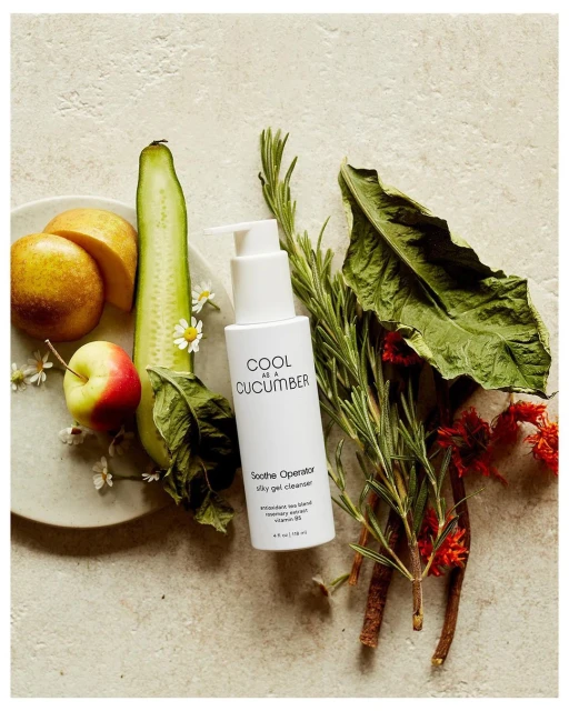 Still life of skincare product bottle with cucumber, rosemary sprigs, apple, and sliced pears on an off-white surface.