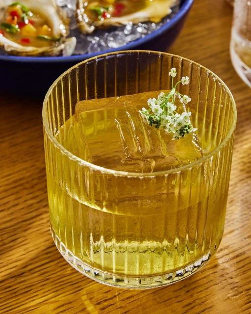 A cocktail in a clear, vertically ribbed glass with a large ice cube, garnished with small white flowers, with oysters on ice in the background.
