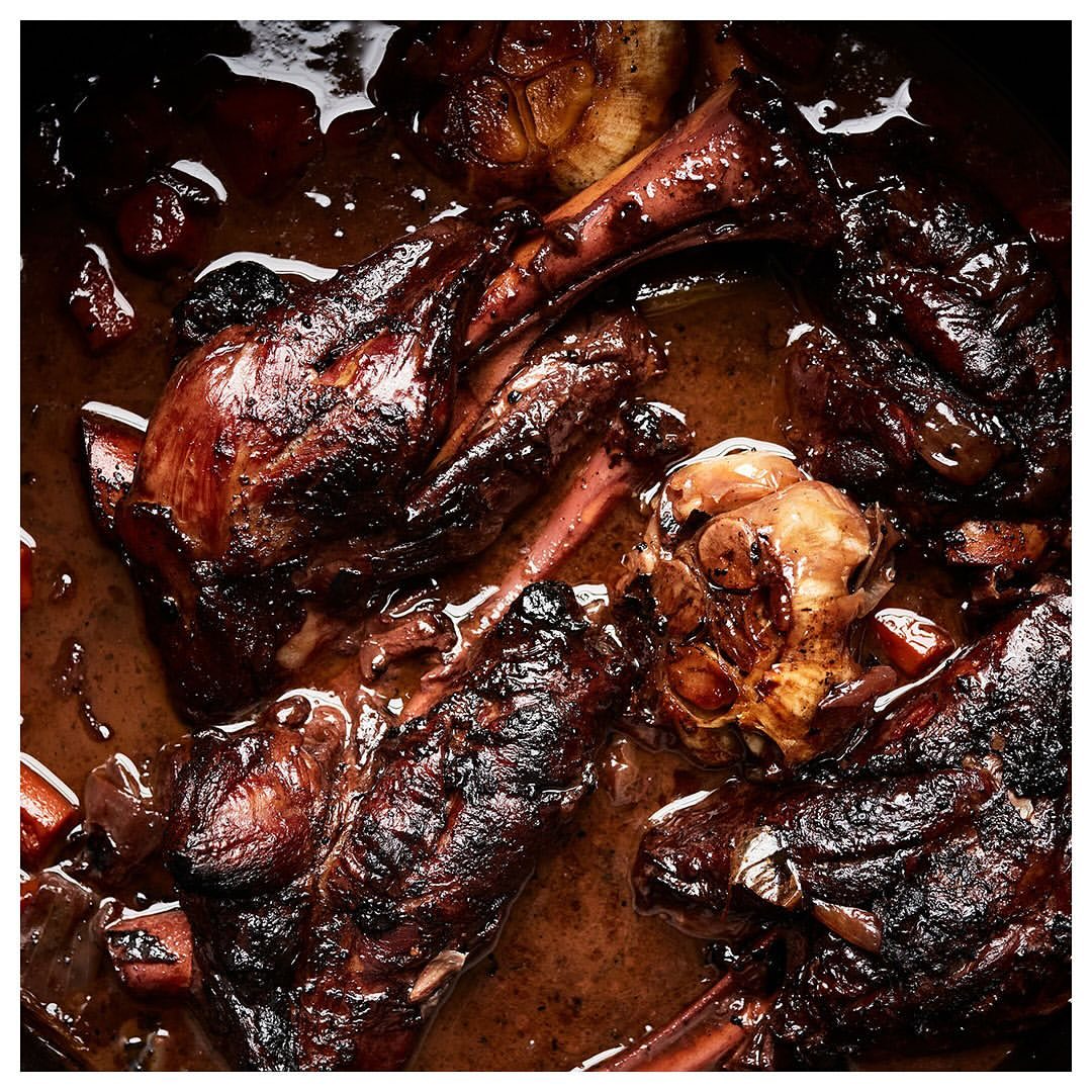 Overhead shot of braised lamb shanks with garlic in a dark, rich sauce, glistening with rendered fat in warm lighting.