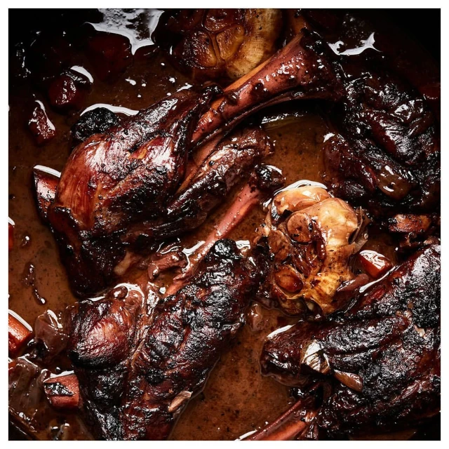 Overhead shot of braised lamb shanks with garlic in a dark, rich sauce, glistening with rendered fat in warm lighting.