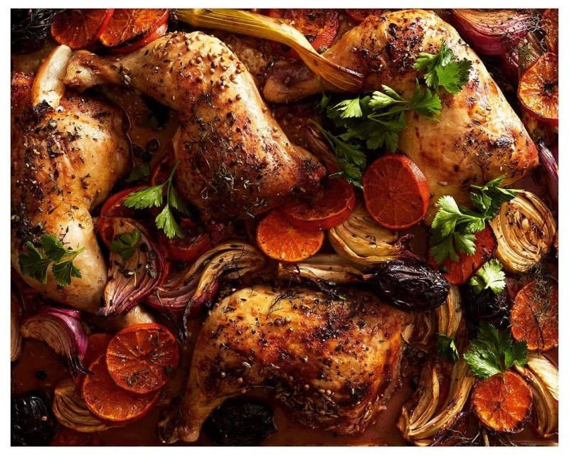 Overhead shot of four roasted chicken legs surrounded by sliced oranges, onions, plums, and herbs, creating a vibrant food still life.