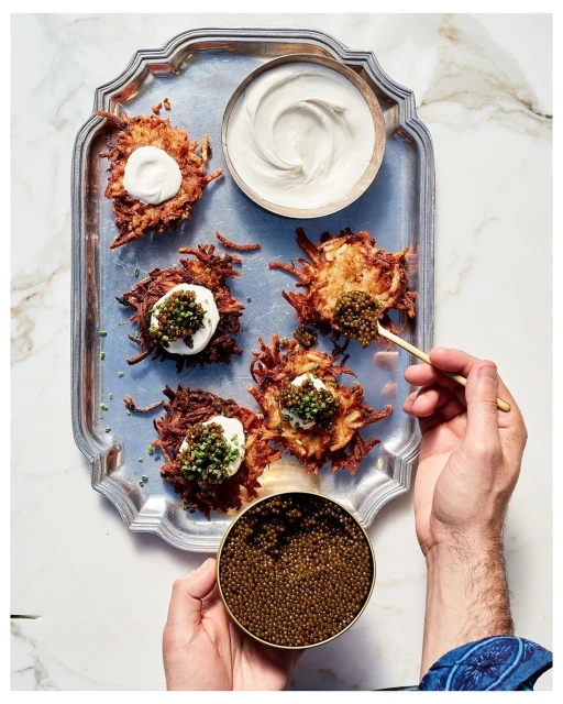 Overhead shot of potato latkes topped with cream and caviar, served on a silver tray, with a human hand holding a caviar tin.