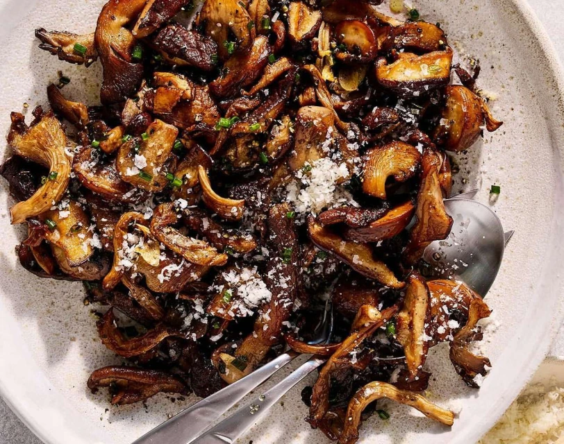 Overhead shot of sauteed mushrooms in a white bowl, garnished with herbs and sprinkled with grated cheese, with a metal serving spoon.