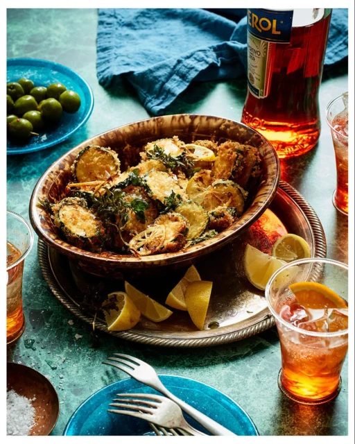 Overhead shot featuring fried zucchini slices in a ceramic bowl, glasses of orange cocktail, and a bottle of Aperol on a marble surface.