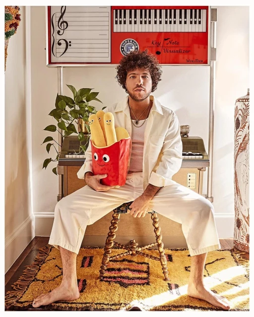 Adult male with curly hair sits on a stool holding a plush french fry toy, surrounded by vintage furniture and warm yellow light.