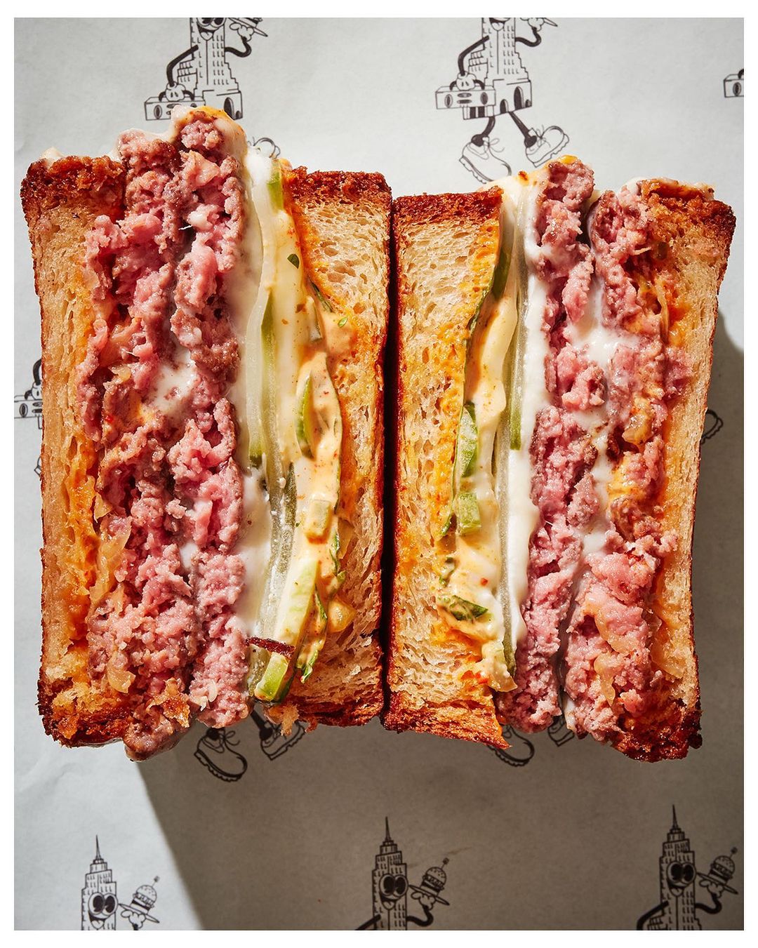 Eye-level, close-up of a halved sandwich featuring meat and vegetables between toasted bread slices on patterned white paper.