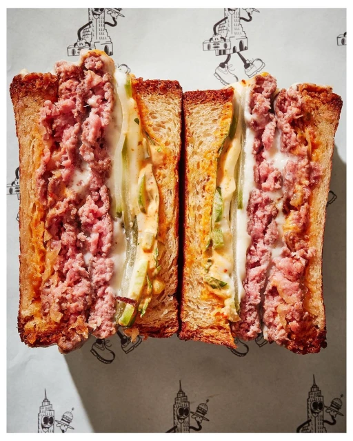 Eye-level, close-up of a halved sandwich featuring meat and vegetables between toasted bread slices on patterned white paper. 
