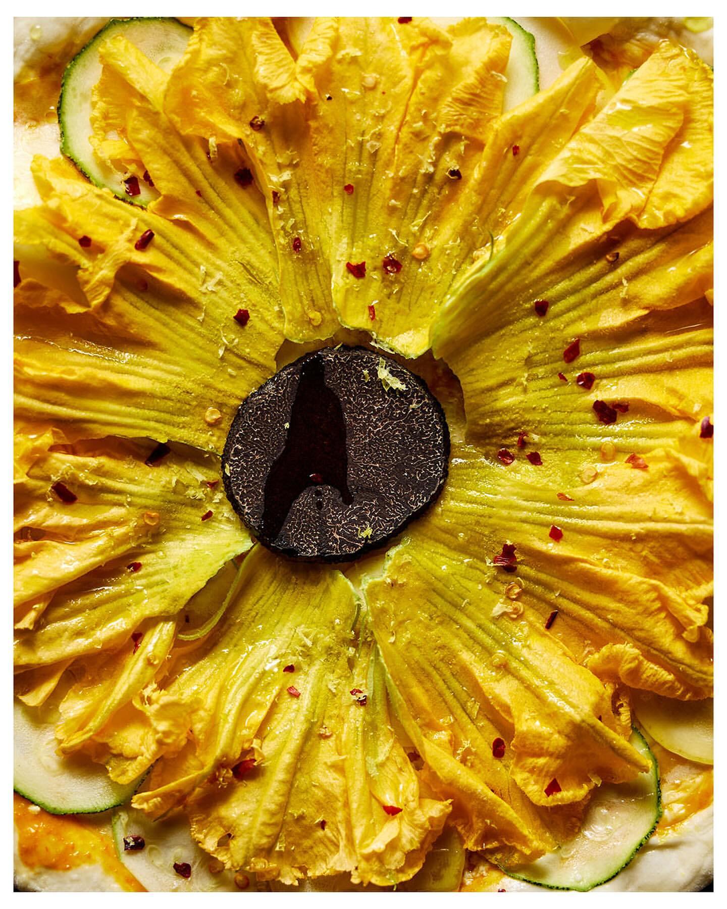 Overhead shot of a pizza featuring yellow squash blossoms, slices of zucchini, truffle, and red pepper flakes on a light-colored crust.