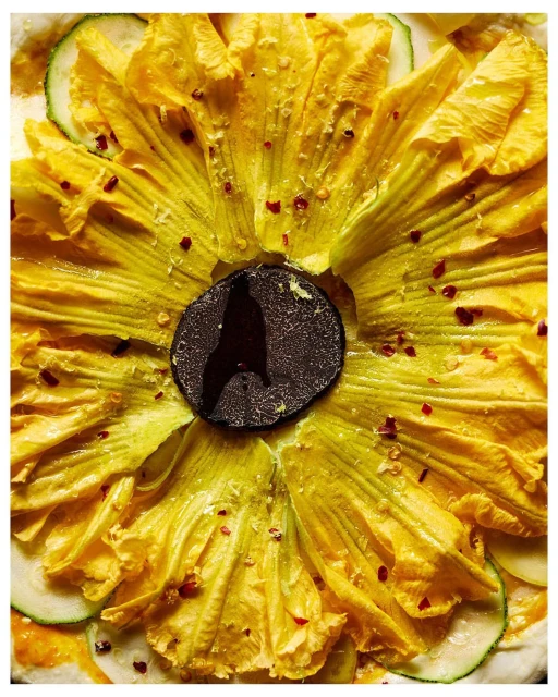 Overhead shot of a pizza featuring yellow squash blossoms, slices of zucchini, truffle, and red pepper flakes on a light-colored crust.