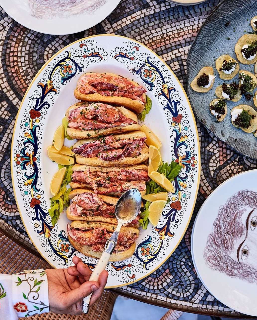 Overhead view of an ornamental platter filled with stacked lobster rolls, garnished with lemon wedges, and set on a mosaic table.
