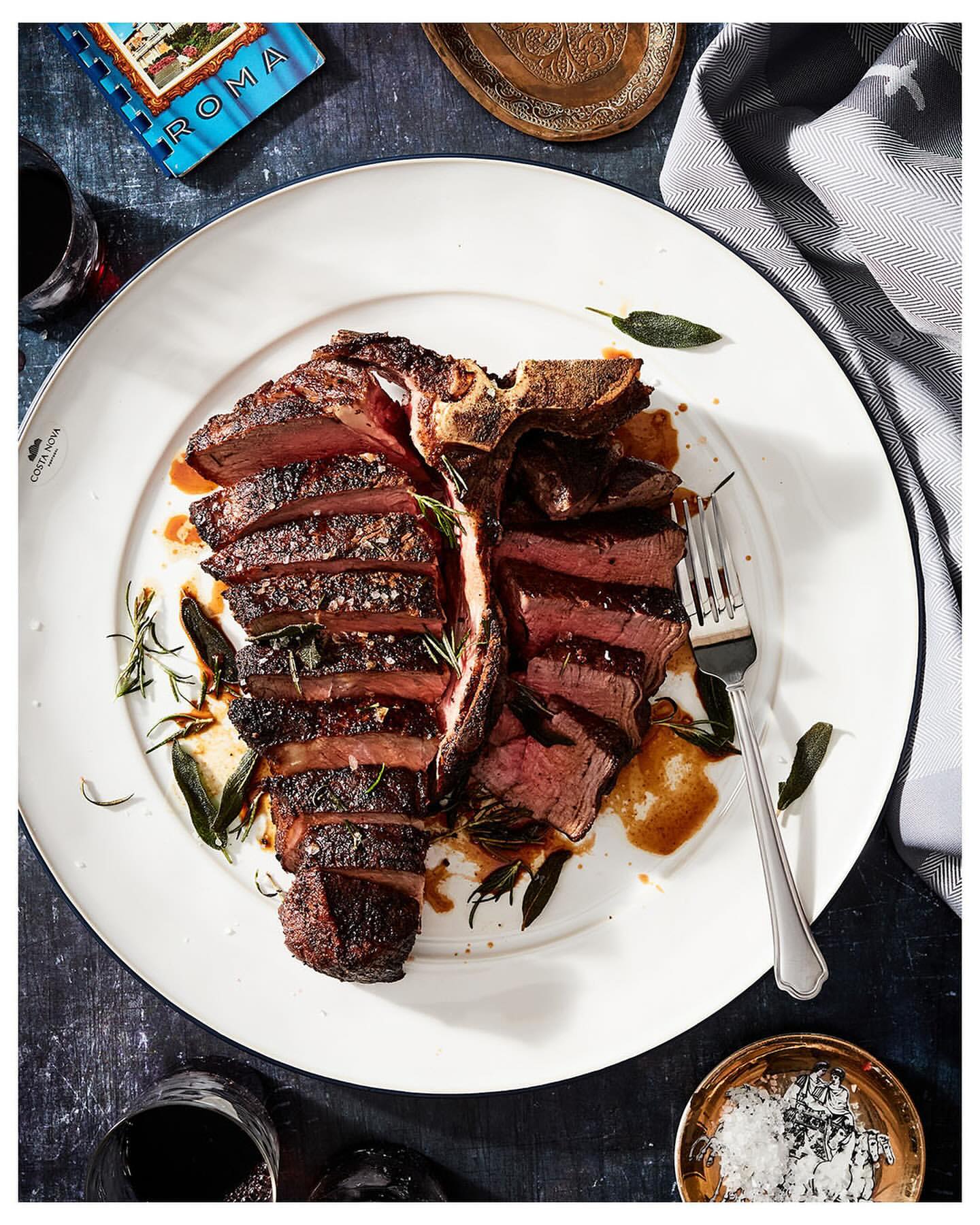 Overhead shot of a sliced T-bone steak on a white plate, garnished with sage and rosemary, alongside red wine and seasonings.