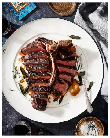 Overhead shot of a sliced T-bone steak on a white plate, garnished with sage and rosemary, alongside red wine and seasonings.