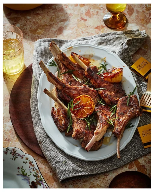 Overhead shot of rack of lamb on a white plate with roasted lemons and rosemary sprigs, complemented by drinks and flatware.