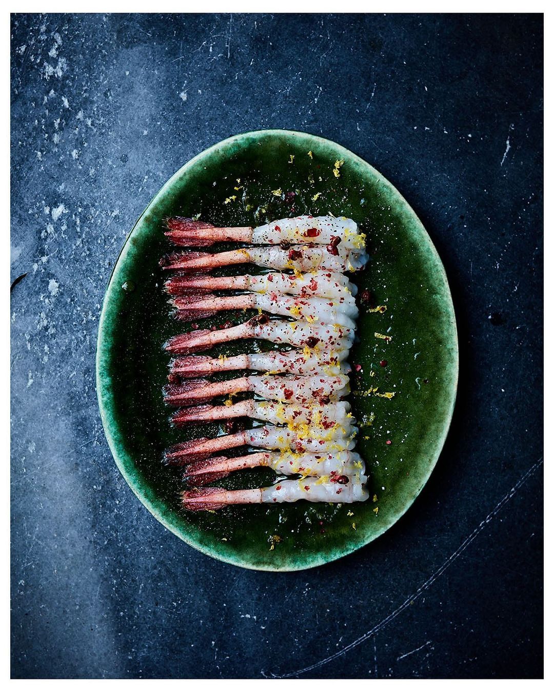 Overhead shot of peeled shrimp on a green plate, seasoned with lemon zest and red pepper flakes, set against a dark slate background.