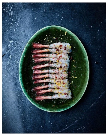 Overhead shot of peeled shrimp on a green plate, seasoned with lemon zest and red pepper flakes, set against a dark slate background.