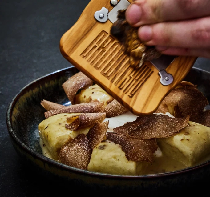 Overhead close-up of a chef grating truffle over gnocchi with cream sauce in a dark bowl against a black surface in a restaurant setting.