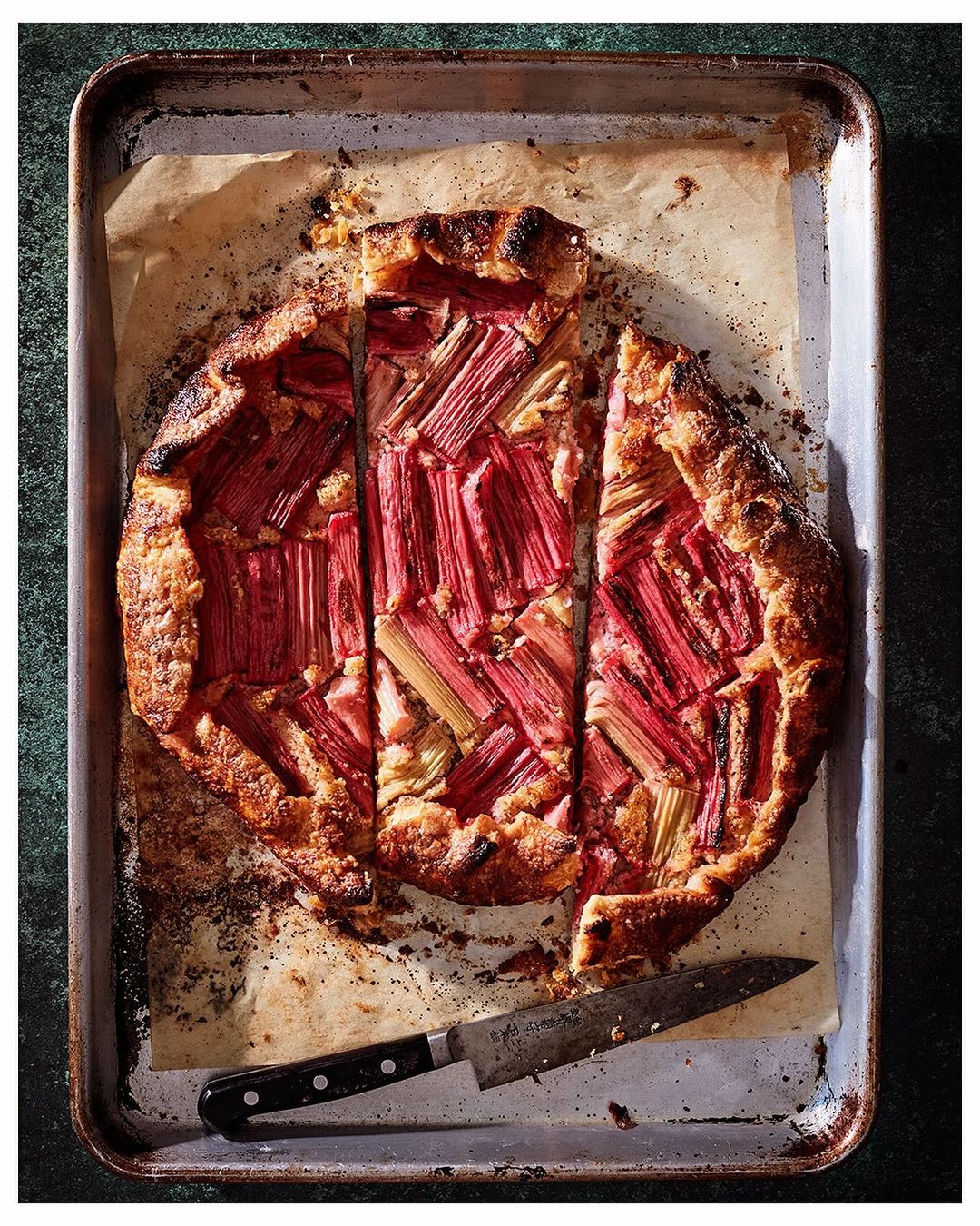 Overhead shot of a baked rhubarb galette with a knife, showcasing the golden crust and pink rhubarb filling on a parchment-lined tray.