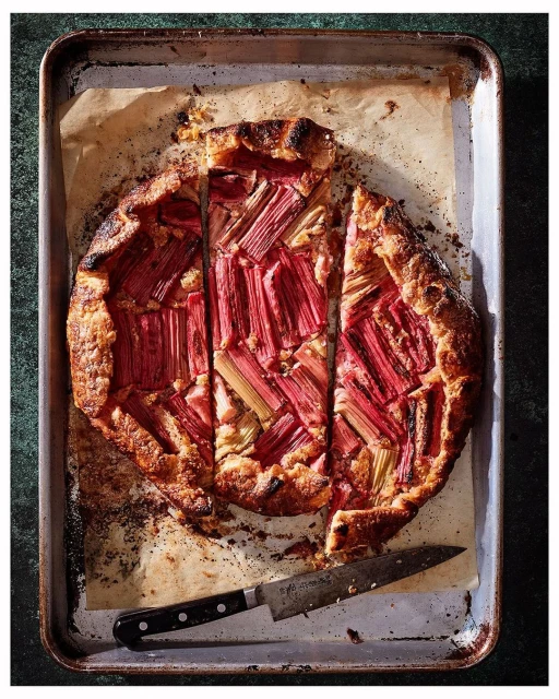 Overhead shot of a baked rhubarb galette with a knife, showcasing the golden crust and pink rhubarb filling on a parchment-lined tray.