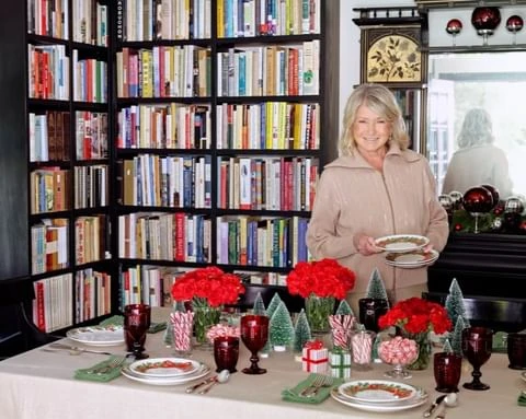 Martha Stewart is standing at a holiday table set with red carnations, small Christmas trees, candy, and decorative plates and glassware.