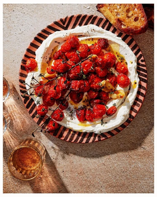 Overhead shot of a patterned plate with roasted tomatoes over creamy white cheese, served with toasted bread and a glass of beverage.