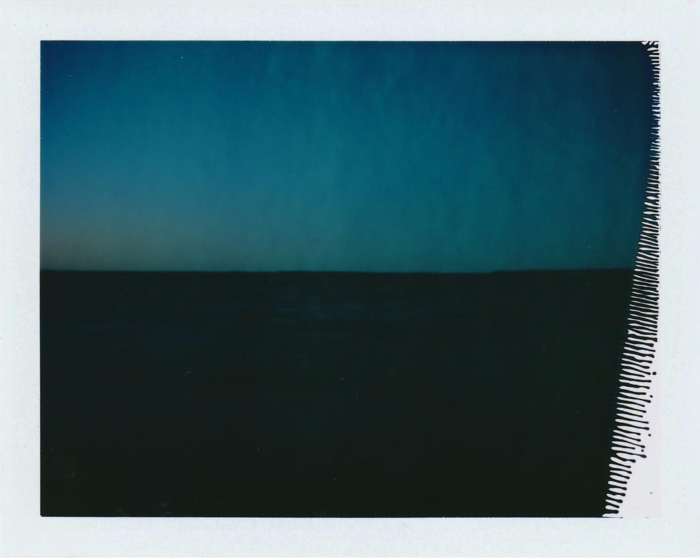 Minimalist landscape featuring a dark, flat field under a gradient of blue sky, captured with a polaroid style and rough texture.