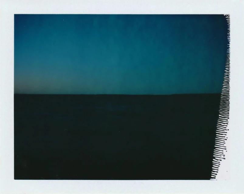 Minimalist landscape featuring a dark, flat field under a gradient of blue sky, captured with a polaroid style and rough texture.