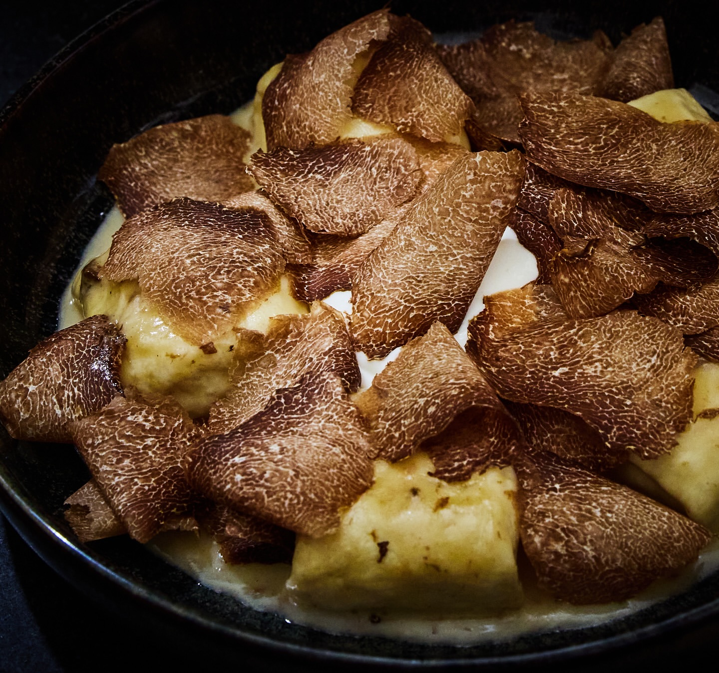 Close-up of gnocchi dish topped with truffle shavings and a light sauce, presented in a black bowl.