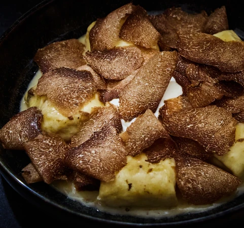 Close-up of gnocchi dish topped with truffle shavings and a light sauce, presented in a black bowl.