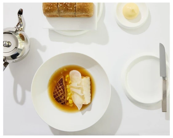 Overhead shot of elegant dish with fish, corn, and broth in white bowl, complemented by bread, butter, silver teapot and knife on white table.