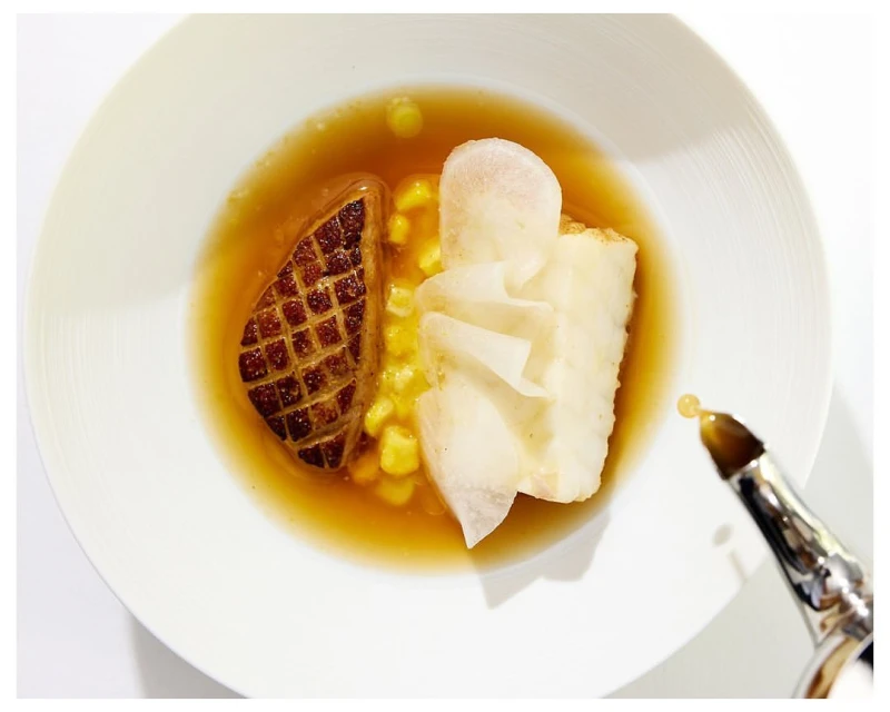 Overhead shot of gourmet plate presentation, featuring foie gras, white asparagus, and broth, drizzled with oil, on a white plate.
