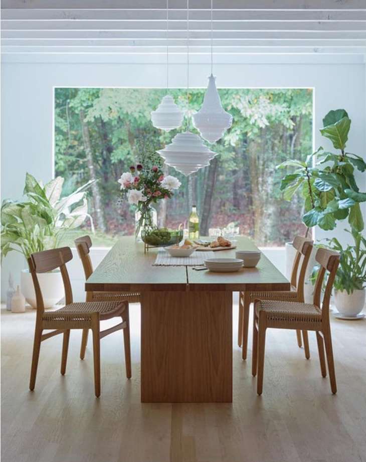 Dining room interior with wooden table set for a meal, surrounded by chairs, and large windows showcasing a lush green outdoor view.