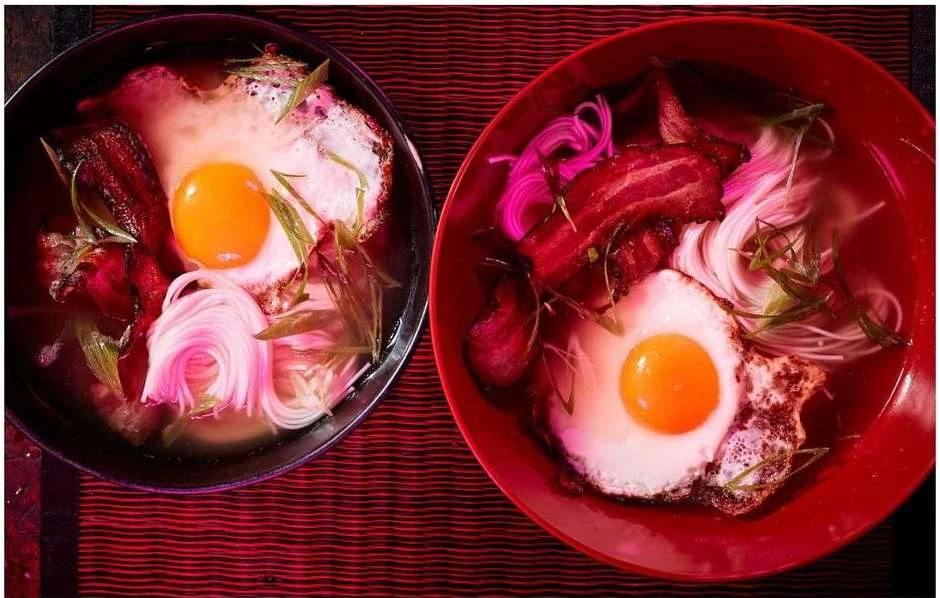 Two bowls of ramen with fried eggs and bacon strips on a patterned red surface, showcasing a delicious food presentation.