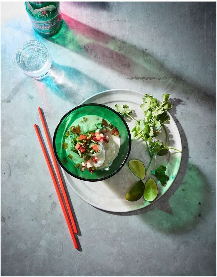 Overhead shot of a green bowl of salsa and white dip with chopsticks, lime, cilantro, and a glass of water on a gray surface.