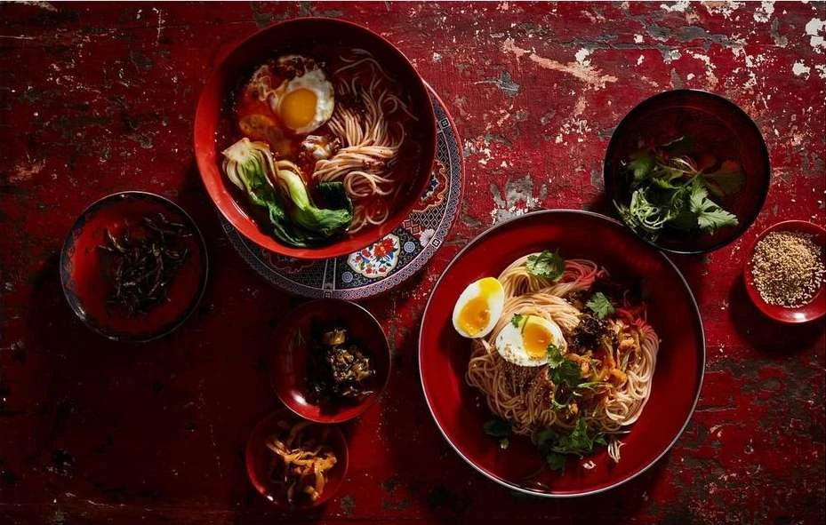 Overhead shot of two bowls of noodle soup with fried and boiled eggs, bok choy, and herbs on a distressed red table with small dishes.