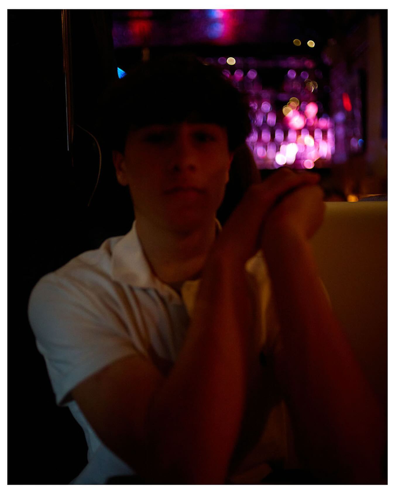 Indoor portrait of a young adult male with dark hair and white collared shirt, sitting in a dimly lit interior with a blurry background.