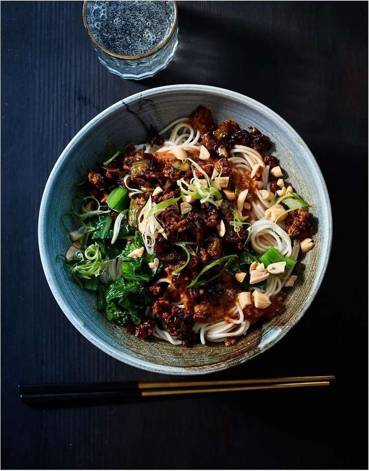 Overhead shot of noodle bowl with meat, greens, and garnish, alongside a glass of water and chopsticks on a dark wood surface.