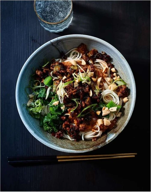 Overhead shot of noodle bowl with meat, greens, and garnish, alongside a glass of water and chopsticks on a dark wood surface.